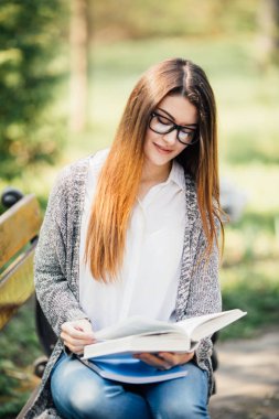 kitap gülümseyen ve kameraya bakarak onun elinde tutan banktaoturuyor, güzel ve mutlu genç öğrenci kız.