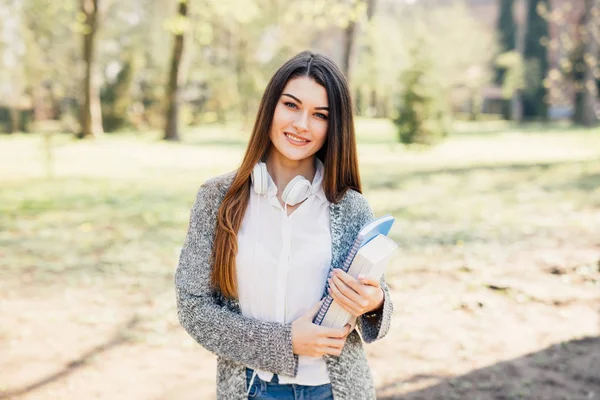 Веселая привлекательная молодая женщина с блокнотами, стоящими и улыбающимися в парке — стоковое фото