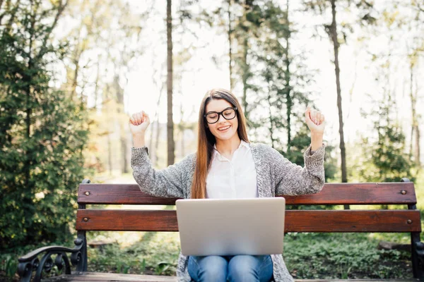 Довольно успешная женщина с поднятыми руками во время работы на ноутбуке, на открытом воздухе в парке скамейка — стоковое фото