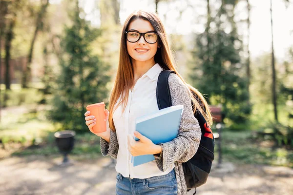 Студентка на улице держит блокнот и улыбается с чашкой кофе — стоковое фото