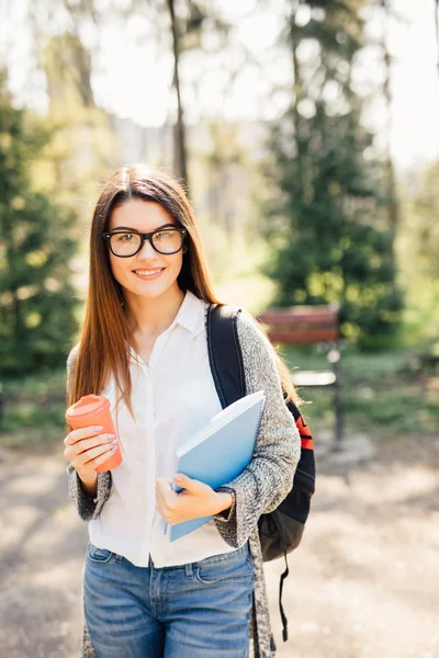 Студентка на улице держит блокнот и улыбается с чашкой кофе — стоковое фото