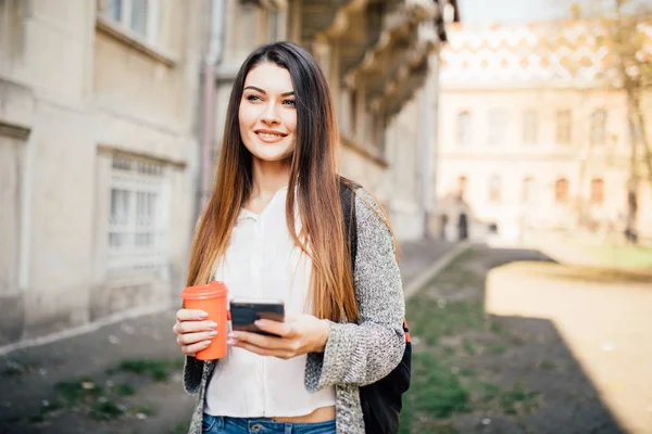 Young woman standing at the street drinking coffee to go and using mobile phone outdoors — Stock Photo, Image