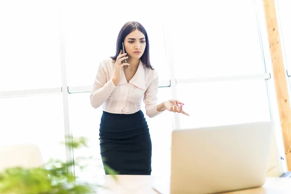 Mujer de negocios hablando por teléfono móvil y mirar el ordenador portátil contra la ventana en la oficina — Foto de Stock