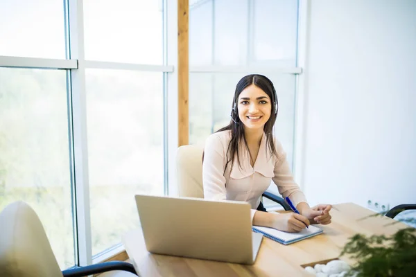 Feliz encantadora joven sentada y trabajando con el ordenador portátil con auriculares en la oficina — Foto de Stock