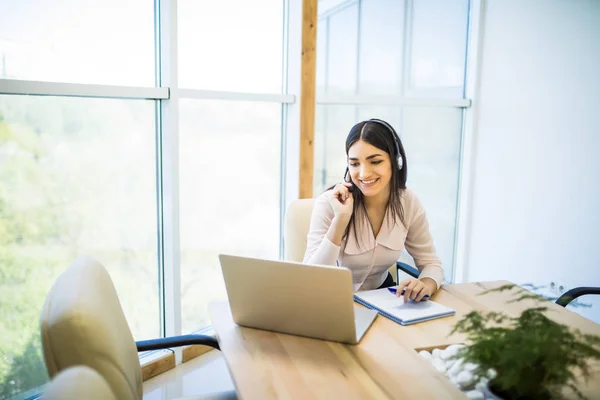 Gelukkig charmant jonge vrouw zitten en werken met laptop met behulp van headset in het kantoor — Stockfoto