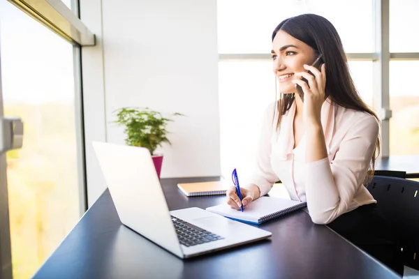 Mujer joven atractiva hablando por teléfono móvil y sonriendo mientras está sentada en su lugar de trabajo en la oficina y mirando a la ventana — Foto de Stock