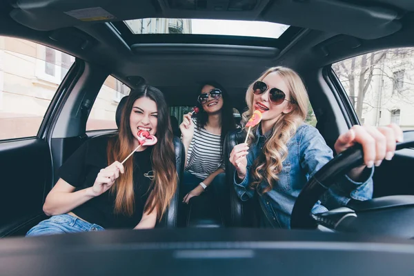 Drie mooie jonge vrolijke vrouwen kijken elkaar met glimlach en lollies eten zittend in de auto — Stockfoto