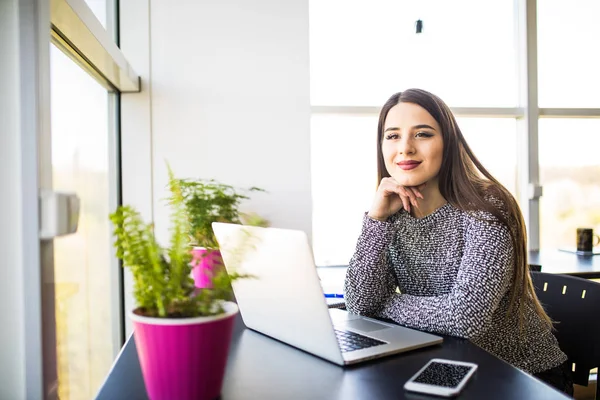 Sorridente giovane donna d'affari seduta alla scrivania dell'ufficio e digitando su un computer portatile, sta guardando la fotocamera — Foto Stock