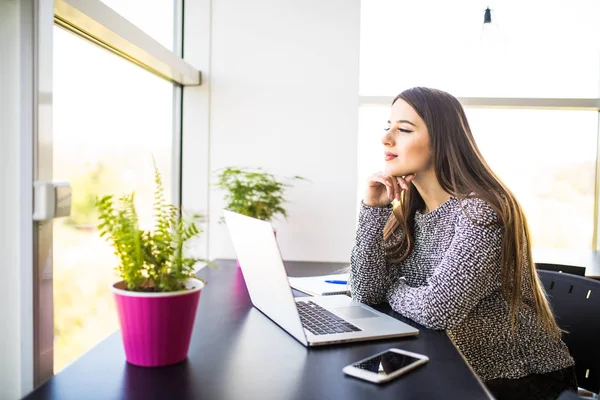 Pensativo joven mujer de negocios hermosa que trabaja en el ordenador portátil y mantener la mano en la barbilla mientras está sentado en su lugar de trabajo — Foto de Stock