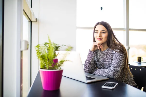 Pensativo joven mujer de negocios hermosa que trabaja en el ordenador portátil y mantener la mano en la barbilla mientras está sentado en su lugar de trabajo — Foto de Stock