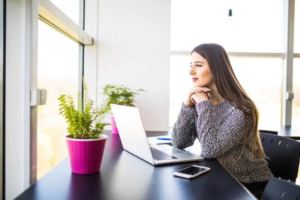 Mujer de oficina bonita pensativa sentada en su escritorio con computadora portátil, mirando a la distancia con la mano en la barbilla . — Foto de Stock