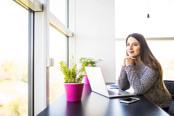 Sonriente joven mujer de negocios sentada en el escritorio de la oficina y escribiendo en un ordenador portátil, ella está mirando a la cámara — Foto de Stock