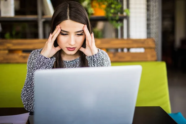 Молода розчарована жінка працює на офісному столі перед ноутбуком, що страждає від хронічних щоденних головних болів — стокове фото