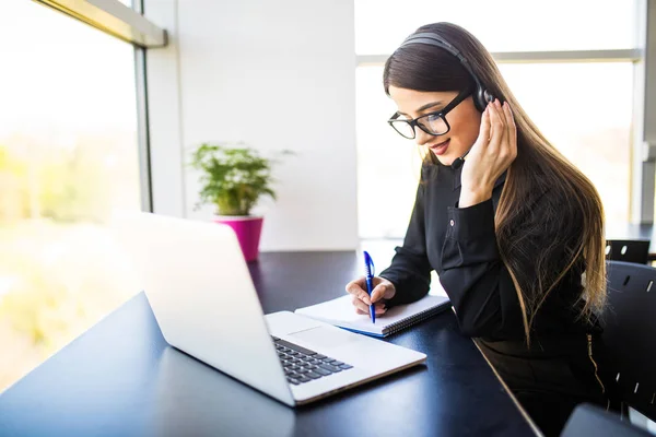 Glad ung kvinna manager tar anteckningar på goda nyheter på headsetet och anteckningsboken i office — Stockfoto