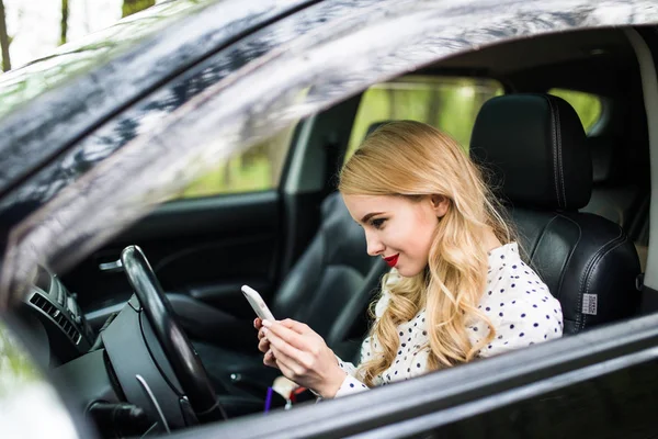 Hermosa mujer está sentada y conduciendo un coche sosteniendo un teléfono móvil y mensajería . — Foto de Stock