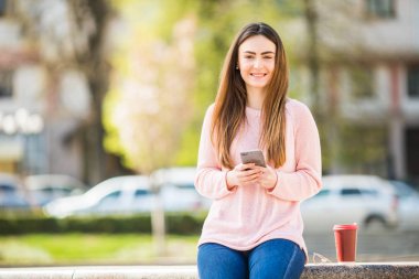 Young woman with phone browse in internet and drink coffee in the city street clipart