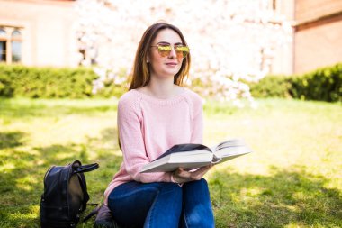 genç, çekici kız öğrenci yeşil çim ve çiçek ağaç çerçevede kitap okur