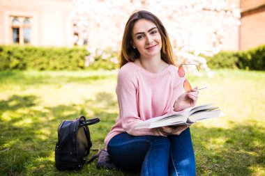 genç, çekici kız öğrenci yeşil çim ve çiçek ağaç çerçevede kitap okur