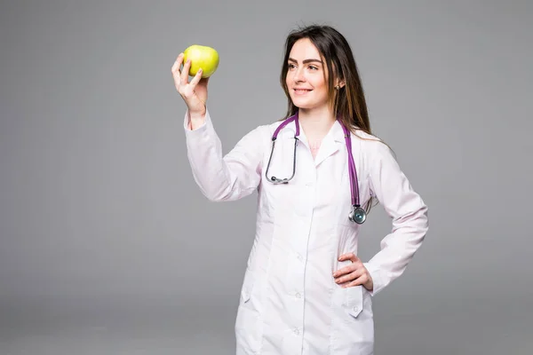 Alegre linda joven médico de pie y sosteniendo una manzana en gris — Foto de Stock