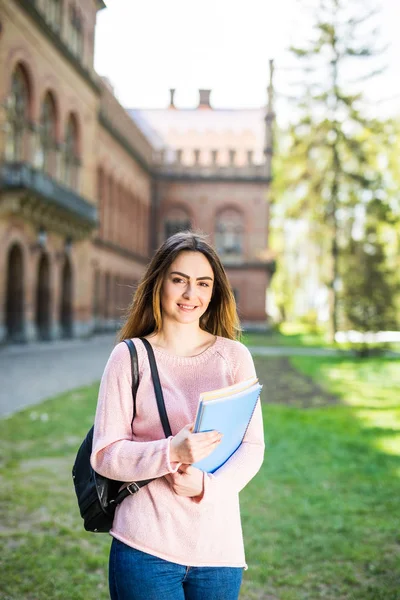 Fröhliche attraktive junge Frau mit Rucksack und Notizbüchern steht und lächelt im Park in der Nähe der Universität — Stockfoto