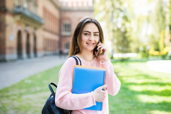 Студентка колледжа с книгами и сумками сидит за пределами университета и говорит по телефону — стоковое фото