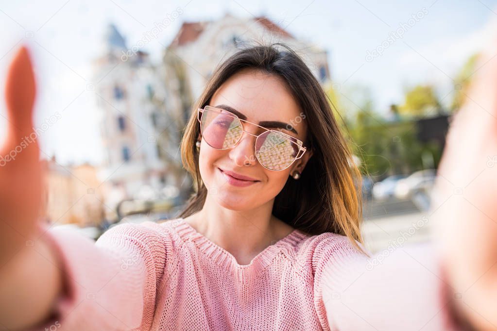 Young woman taking selfie from hands with mobile phone in sunny city street.
