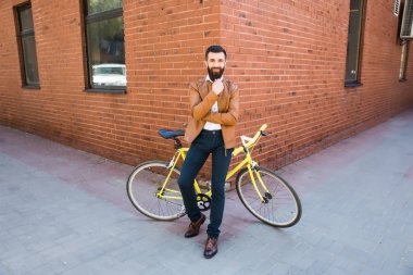 Çeneme elini tutarak ve Onun Bisiklet açık havada yakınındaki duvara yakın otururken kameraya bakarak adam genç sakallı