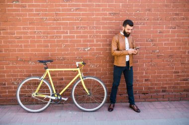 Genç hipster adam onun cellphone tuğla duvar üzerinde sabit bisiklet ile kontrol