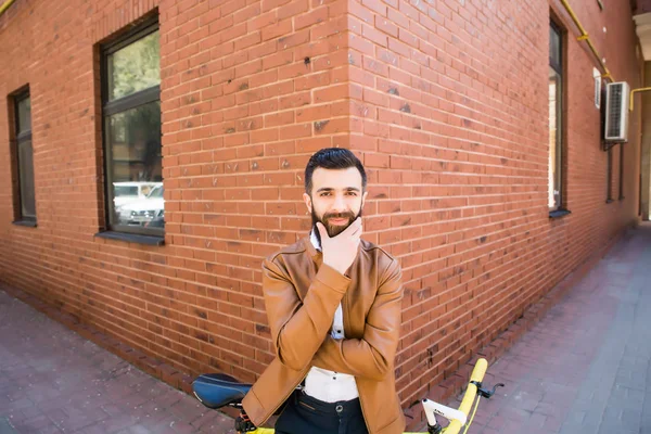 Homem barbudo jovem segurando a mão no queixo e olhando para a câmera enquanto sentado perto de sua bicicleta ao ar livre perto da parede de tijolo — Fotografia de Stock