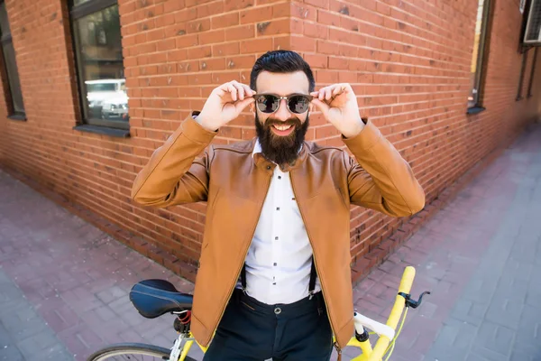 レンガの背景に自転車の上に座ってサングラスのひげを持つスタイリッシュな若者 — ストック写真