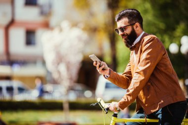 Yakışıklı sakallı hippi adam akıllı bir telefon kullanarak ve şehirde bisiklet sürme sırasında gülümseyen
