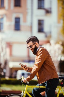 Yakışıklı sakallı hippi adam akıllı bir telefon kullanarak ve şehirde bisiklet sürme sırasında gülümseyen