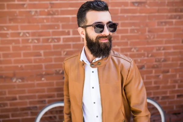 Retrato de cerca de un hombre barbudo brutal sobre fondo de pared de ladrillo. Joven hipster elegante posando en la calle . — Foto de Stock