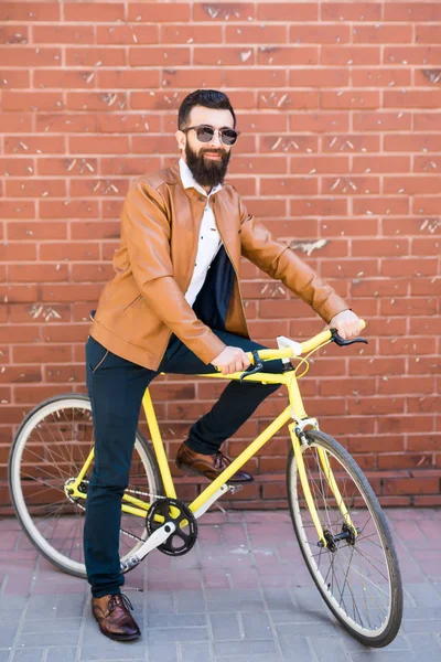 Homem bonito jovem com uma barba contra a parede de tijolo senta-se em uma bicicleta — Fotografia de Stock
