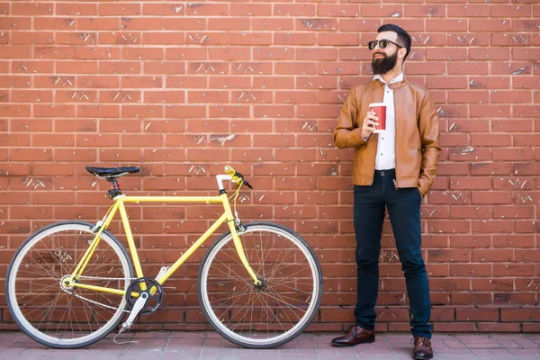 Junger bärtiger Mann trinkt Kaffee, während er im Freien neben seinem Fahrrad sitzt — Stockfoto