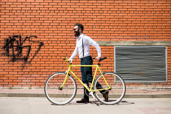 Jovem homem barbudo elegante vai trabalhar de bicicleta contra parede de tijolo — Fotografia de Stock