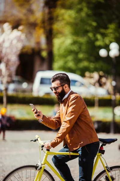 Bonito homem hipster barbudo está usando um telefone inteligente e sorrindo ao andar de bicicleta na cidade — Fotografia de Stock