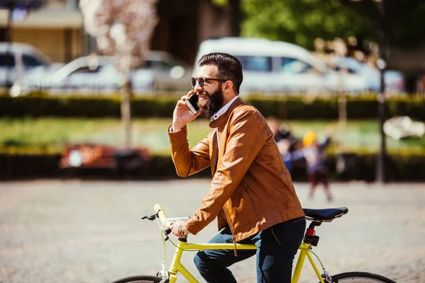 Jovem alegre com barba falando no celular e sorrindo perto de sua bicicleta . — Fotografia de Stock