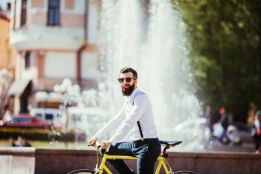 Güneş gözlüğü, sakallı hippi genç adam binmek bisiklet yakınındaki çeşmeler ile şehir.
