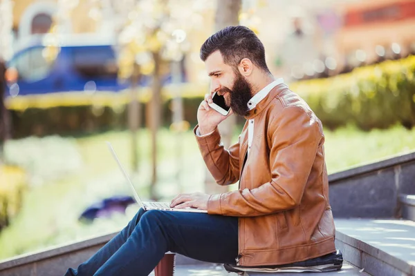 Hipster όμορφος νεαρός άνδρας κάθεται δίπλα του φορητού υπολογιστή στις σκάλες, ενώ μιλώντας στο τηλέφωνό του — Φωτογραφία Αρχείου