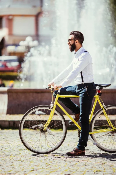 Güneş gözlüğü, sakallı hippi genç adam binmek bisiklet yakınındaki çeşmeler ile şehir. — Stok fotoğraf