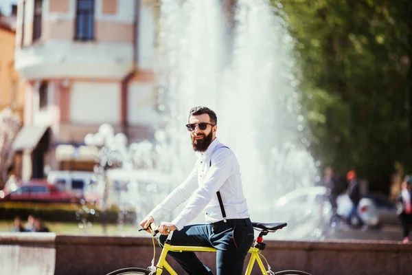 Hipster jovem com uma barba em óculos de sol, andar na cidade com bicicleta perto de fontes . — Fotografia de Stock