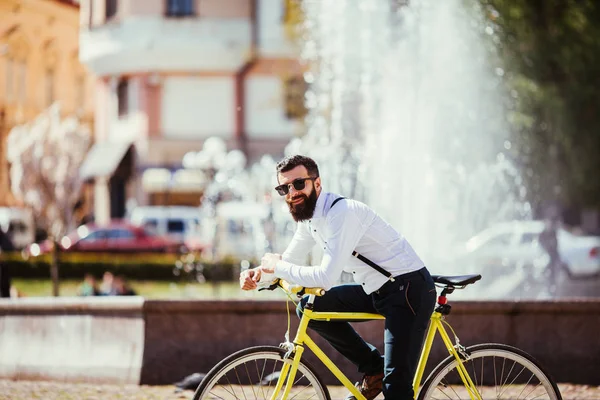 Bonito hipster jovem barbudo homem olhando para a câmera enquanto sentado em sua bicicleta ao ar livre perto de fontes — Fotografia de Stock