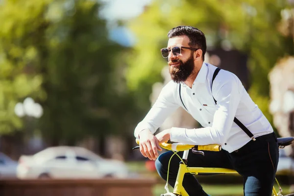 Молодий бородатий чоловік хіпстер в сонцезахисних окулярах, спираючись на свій велосипед і дивлячись далеко, стоячи на міській вулиці — стокове фото