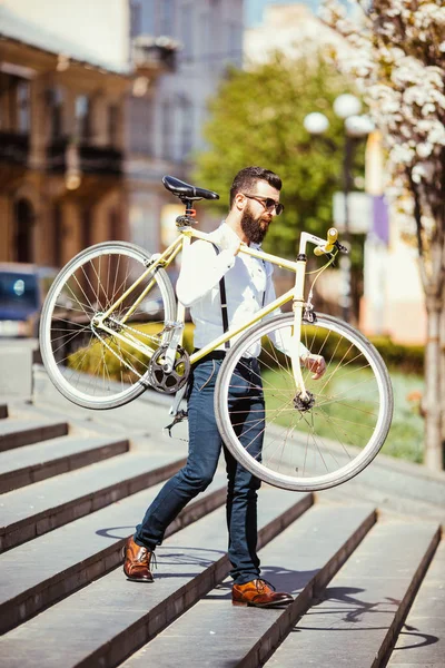 Hipster adam omuz bisikletiyle sokak merdiven üzerinde taşıyan güneş gözlüğü. Bisiklet sağ kolu üzerinde tutarak sakallı yakışıklı bir genç adam. — Stok fotoğraf