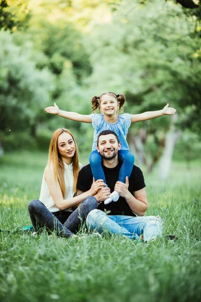 Jeune famille heureuse de trois personnes s'amusant ensemble en plein air. Jolie petite fille sur le dos de son père avec les mains levées heureuses. Parents et fille ont l'air heureux et sourire . — Photo