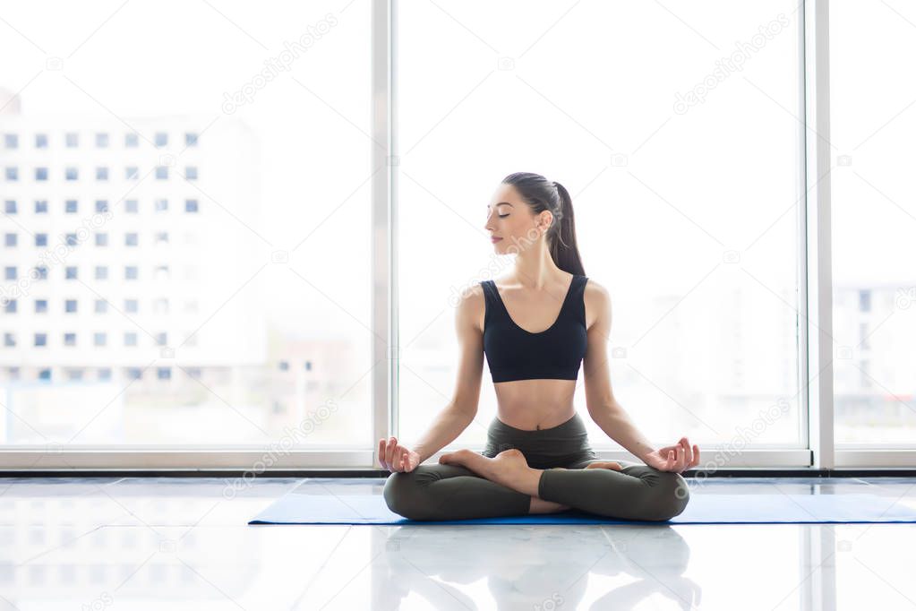 Meditation lotus pose. Young attractive woman practicing yoga, sitting in exercise, working out, wearing sportswear at floor window with city view