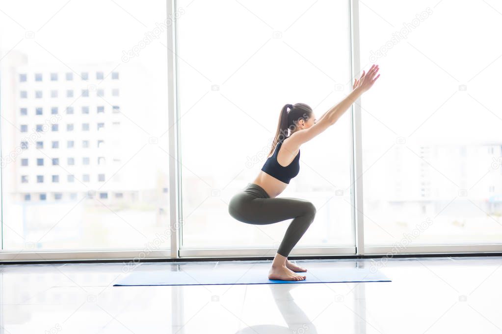 Awkward. Young attractive woman practicing yoga, standing yoga exercise, wearing sportswear near floor window with city view