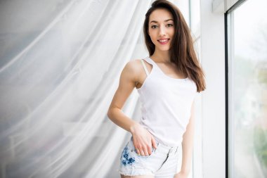 Pencerenin perdelerini açan mutlu bir kadının yan görüntüsü ve dışarıdan gelen sıcak bir ışıkla yeni bir günün tadını çıkarıyor.