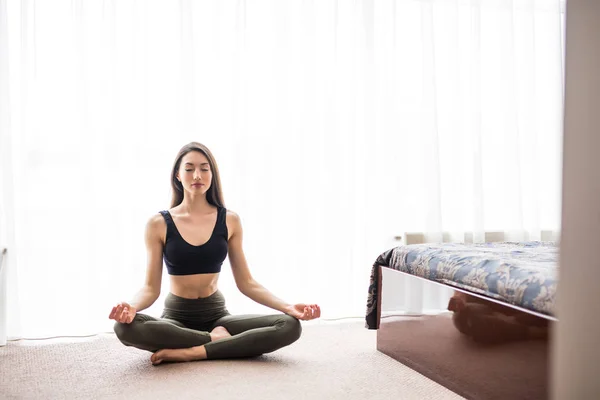 Atractiva joven mujer haciendo ejercicio y sentada en posición de loto de yoga mientras descansa en casa —  Fotos de Stock